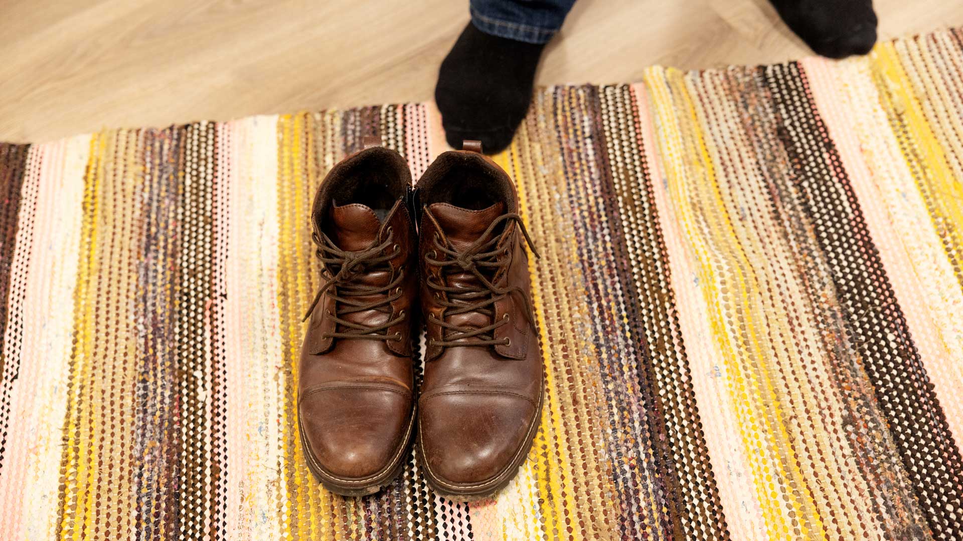 Kuvassa miesten varsikengät räsymatolla. Taustalla näkyy jalat sukkasillaan.