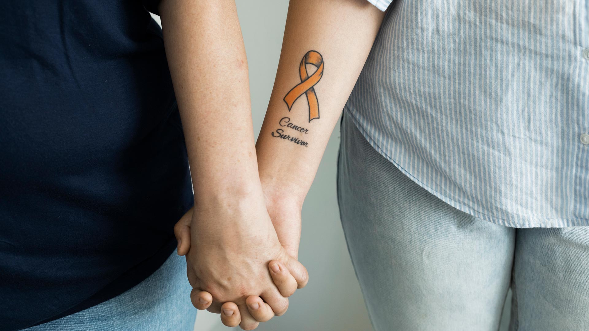 Lähikuva henkilöistä jotka pitävät toisiaan käsistä. Toisen käsivarteen on tatuoitu teksti Cancer Survivor.